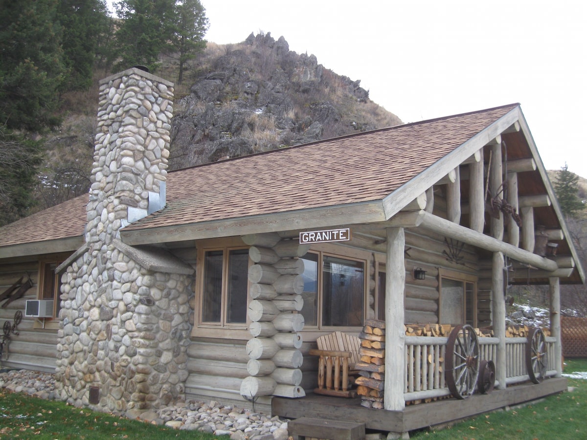 The exterior of a rustic log cabin is showcased, featuring a prominent stone chimney and wooden accents. A mountain backdrop rises in the distance, while neatly stacked firewood adds to the cabin's charm. A sign reading 'Granite' is visible above the entrance.