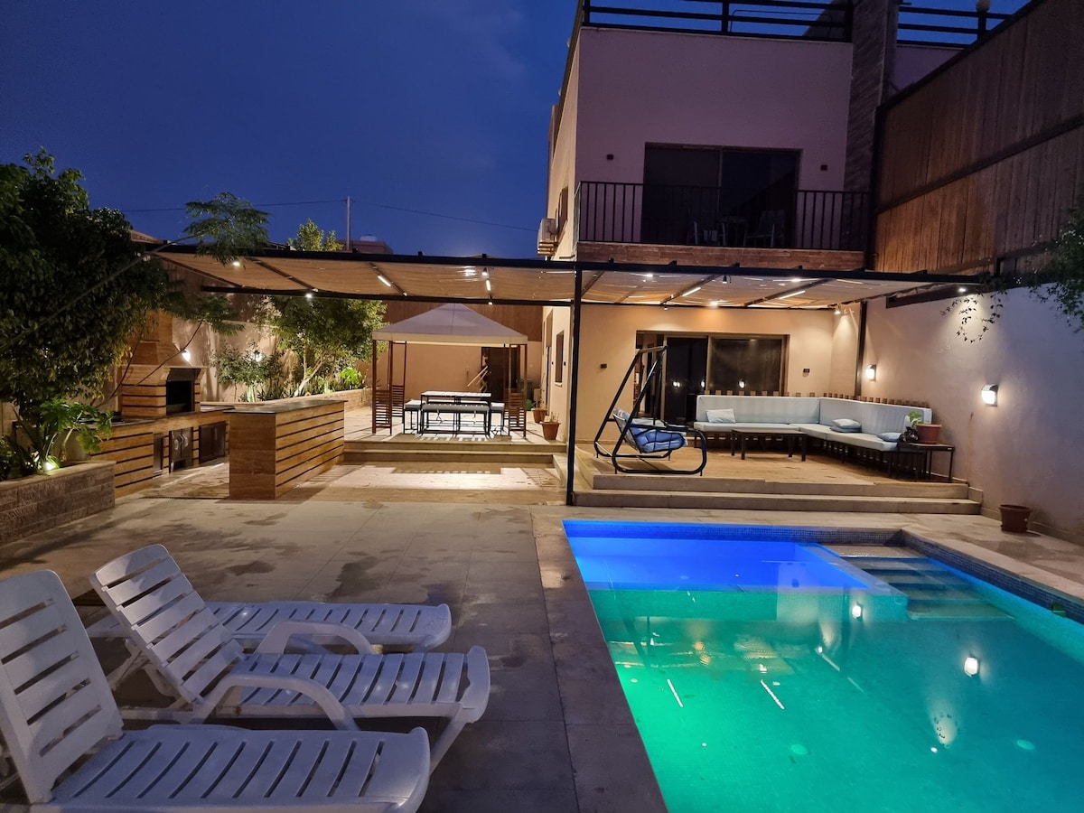 An outdoor area is presented with a heated swimming pool reflecting soft lights. Sun loungers are arranged nearby. A covered patio features a seating area, a dining table, and a swing chair, surrounded by lush greenery, creating a relaxing environment.