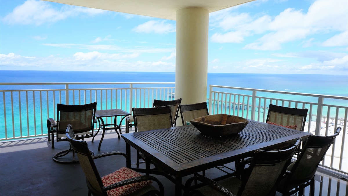 A spacious balcony offers a large dining table surrounded by six chairs, with stunning ocean views visible beyond the railing. The scene is complemented by a decorative bowl at the table's center, and the bright blue sky enhances the tranquil coastal atmosphere.