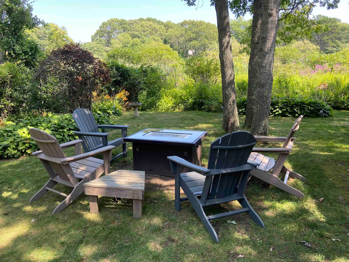 A serene backyard space features five wooden Adirondack chairs arranged around a central fire pit. Lush greenery and flowering plants provide a natural backdrop, creating an inviting setting for outdoor gatherings.