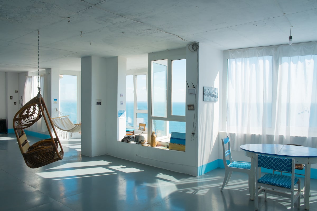 An open-plan living area is filled with natural light, featuring a hanging chair prominently positioned. Large windows offer a direct view of the sea, while a simple dining table is set against a backdrop of white curtains gently billowing in the breeze.
