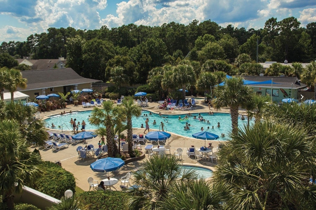 A spacious outdoor pool area is surrounded by lush greenery and palm trees. Numerous lounge chairs and umbrellas are arranged around the pool, where guests enjoy the water. The scene captures both relaxation and recreation under a partly cloudy sky.