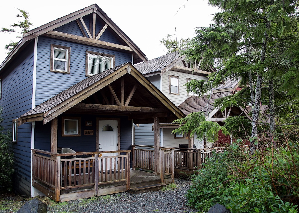 A rustic cabin stands with a blue exterior featuring wooden accents and a covered porch. The entrance is framed by natural foliage, while the surrounding area includes additional cabins and gravel walkways, contributing to a welcoming, forested environment.