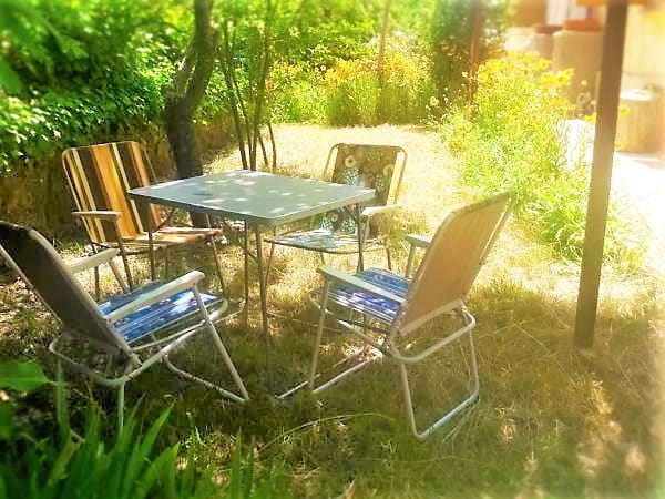 A small outdoor seating area is visible, featuring four chairs arranged around a table. Lush greenery and blooming flowers surround the space, creating a natural setting for relaxation. Sunlight filters through the leaves, casting gentle shadows on the ground.