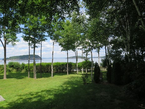 A view of the serene landscape is presented, showcasing a well-maintained lawn bordered by green trees. In the distance, Frenchman Bay is visible, framed by lush foliage, providing an expansive natural backdrop.
