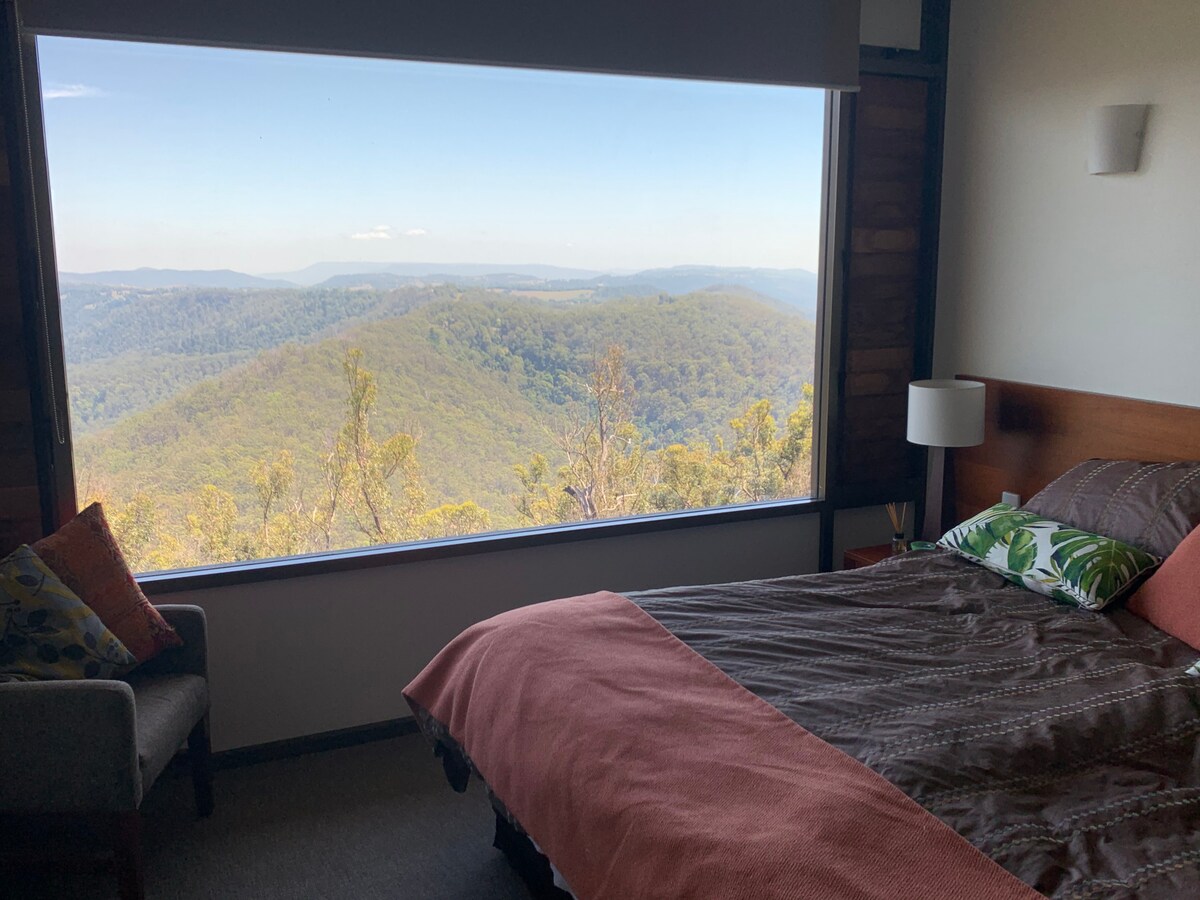 A bedroom features a spacious bed with earthy-toned bedding and decorative pillows. A large window offers expansive views of the surrounding mountains, framed by natural greenery. A cozy armchair sits beside the bed, enhancing the serene atmosphere.