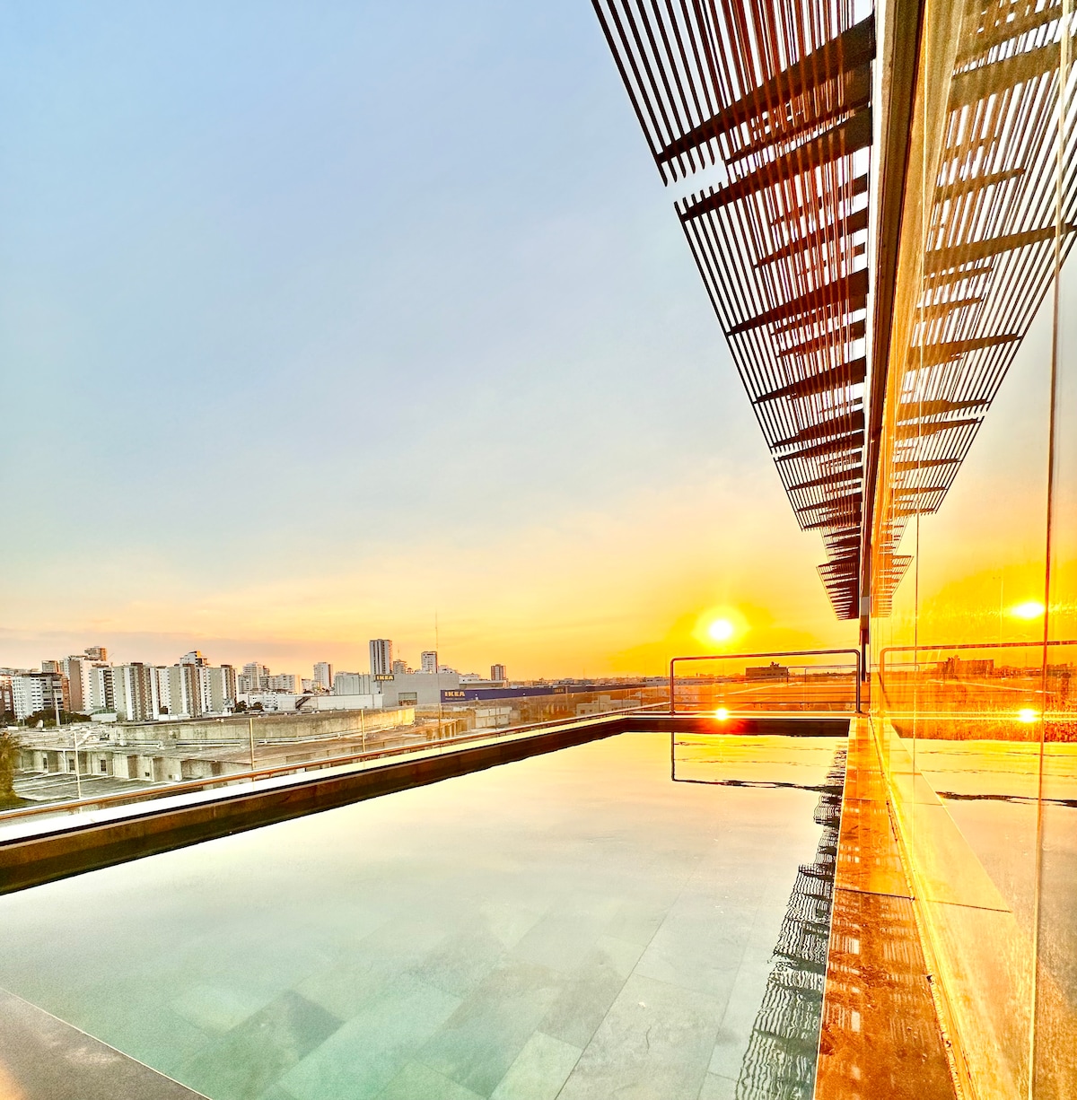 A rooftop terrace is captured during sunset, featuring a shimmering pool that reflects the vibrant colors of the sky. A modern structure with a slatted overhang is visible, framing the scene while providing shade. The skyline of Santo Domingo is outlined in the background.