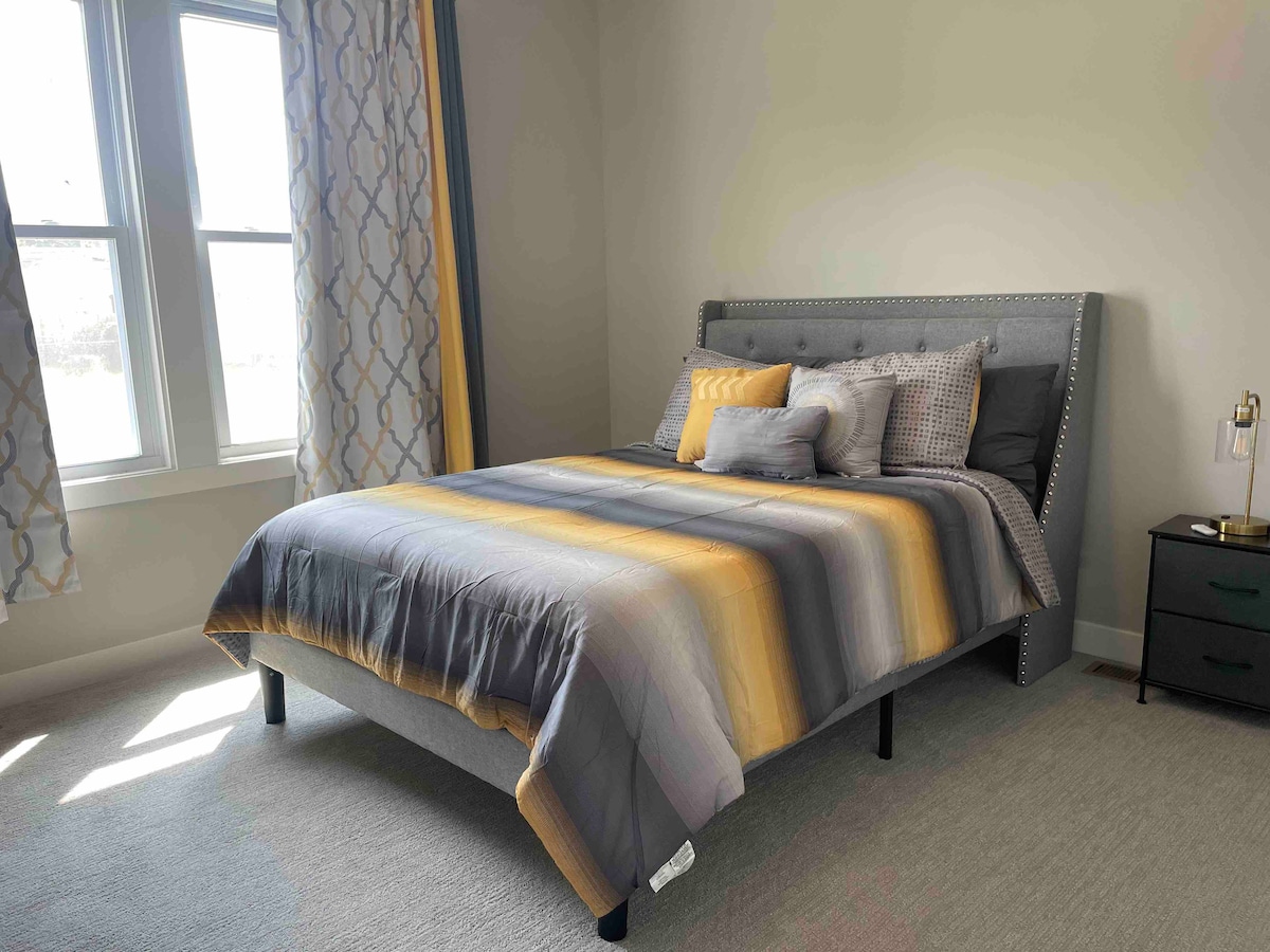 A spacious bed is dressed in a striped grey and yellow comforter, accompanied by various cushions. Sunlight enters through two large windows, illuminating the room. A bedside table is positioned beside the bed, enhancing the overall layout.