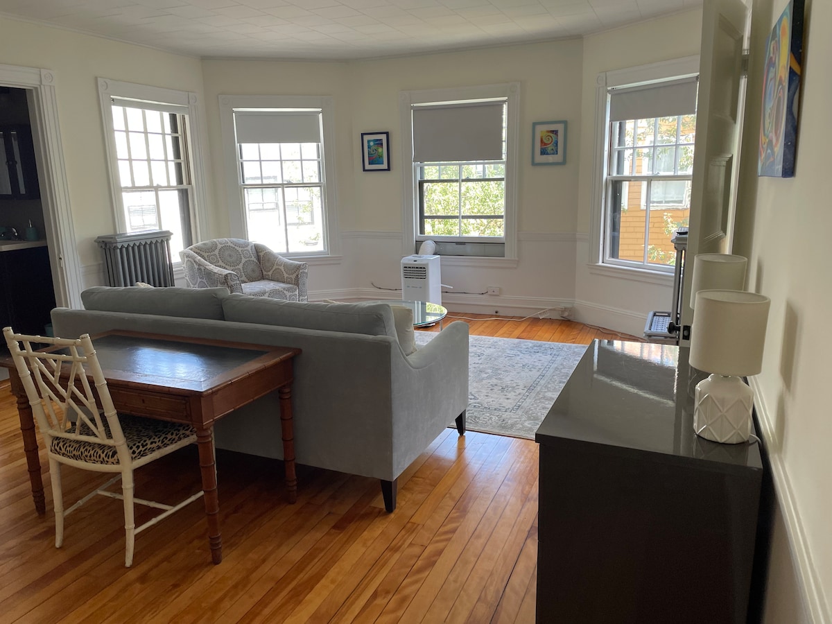 A bright open living space features large windows allowing natural light to fill the room. A cozy seating arrangement is created by a light gray sofa and patterned chair, complemented by a coffee table and a small dining area with a wooden table. Radiator and art pieces add character.