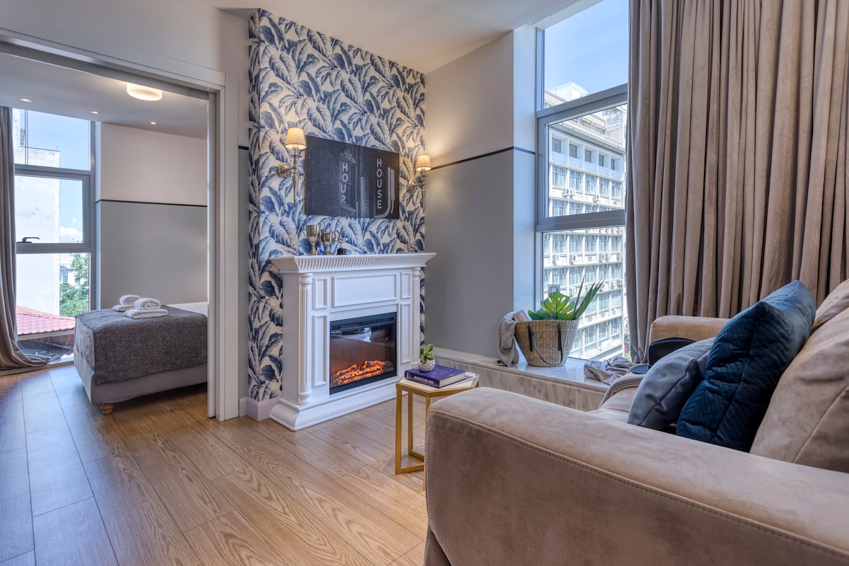 A well-appointed living space features a decorative fireplace, surrounded by elegant furnishings and soft, natural light flowing through floor-to-ceiling windows. A glimpse of the serene bedroom is visible, complemented by charming wallpaper and lush indoor plants.