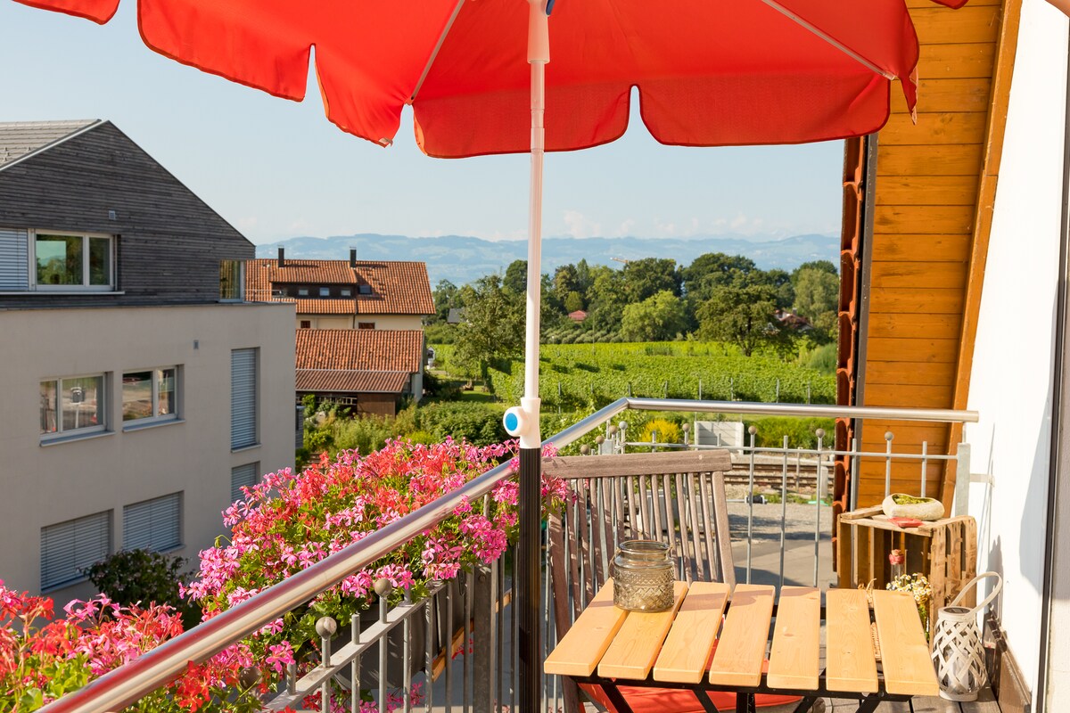A sunlit balcony is enclosed by a vibrant red umbrella, providing shade over a small wooden table and chairs. Potted flowers add color to the railing, while a scenic view of vineyards and distant mountains is visible in the background.
