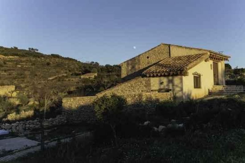 A rustic stone house is situated amidst gentle hills, with elements of traditional architecture visible, including a sloped roof and wooden details. The tranquil environment features lush greenery and a clear sky, suggesting a peaceful rural setting.
