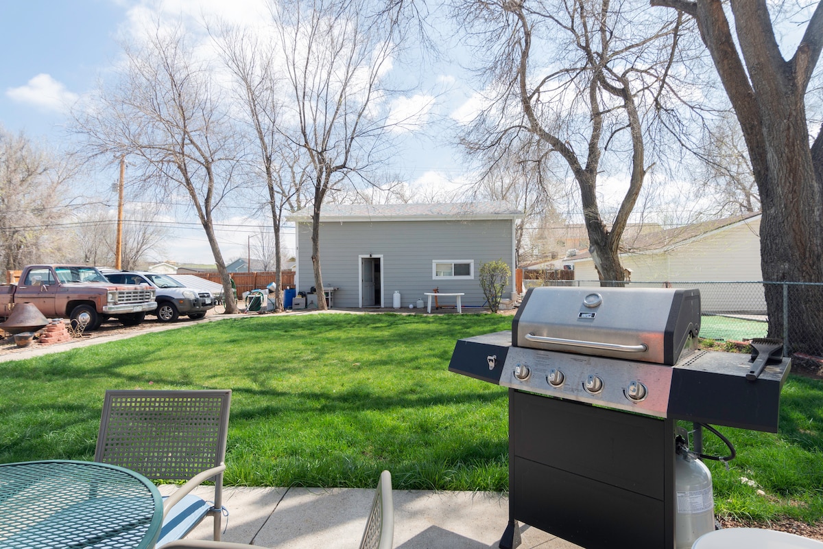 A spacious outdoor area is depicted, featuring a BBQ grill positioned on a stone patio. Lush green grass extends in the background, with a view of a serene yard and nearby trees. A light-colored building is visible, alongside parked vehicles.