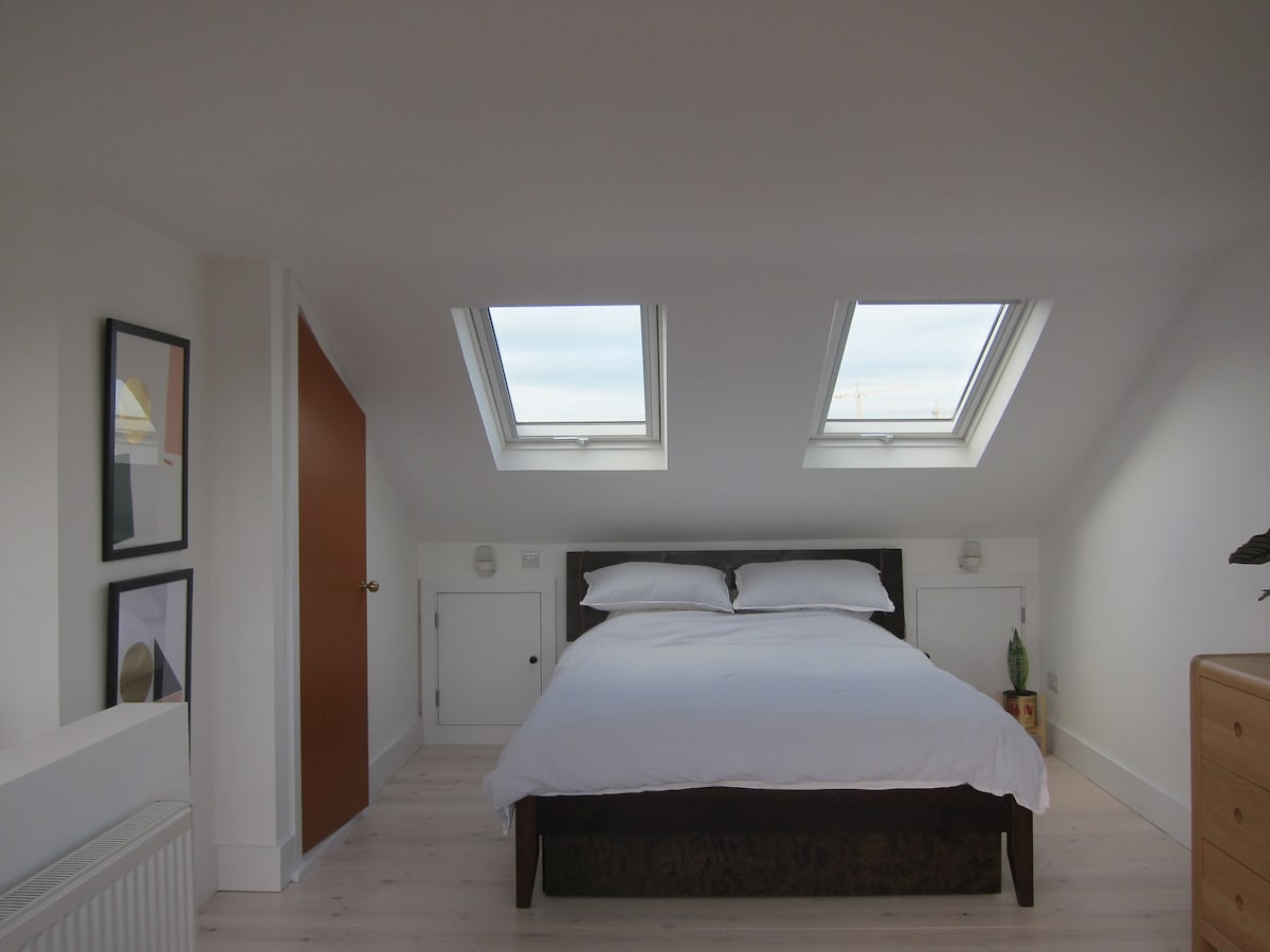 A bright and airy bedroom features a spacious bed with crisp white linens. Two skylights allow natural light to fill the room, while simple artwork adorns the walls. A wooden dresser and a small plant add a touch of warmth to the minimalist decor.