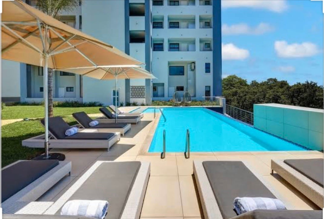 A sunlit pool area features a spacious swimming pool flanked by lounge chairs and umbrellas. Lush greenery surrounds the space, and modern architecture is visible in the background, enhancing the contemporary design of the setting.