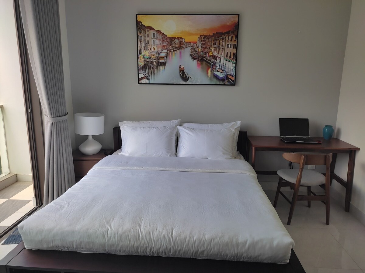 A spacious bed is centered in the room, adorned with fresh white linens. A vibrant landscape painting hangs above, depicting a serene canal scene. To one side, a small desk is paired with a wooden chair, while a bedside lamp provides additional lighting.