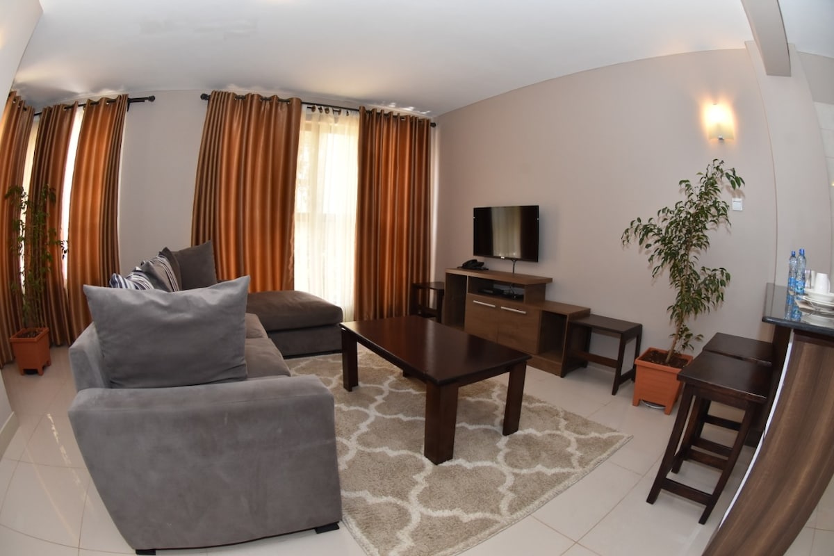 A modern living area features a plush gray sectional sofa with accent pillows, accompanied by a dark wooden coffee table. A flat-screen TV is positioned on a media console against a neutral wall, while large windows dressed in warm curtains allow natural light to filter in.