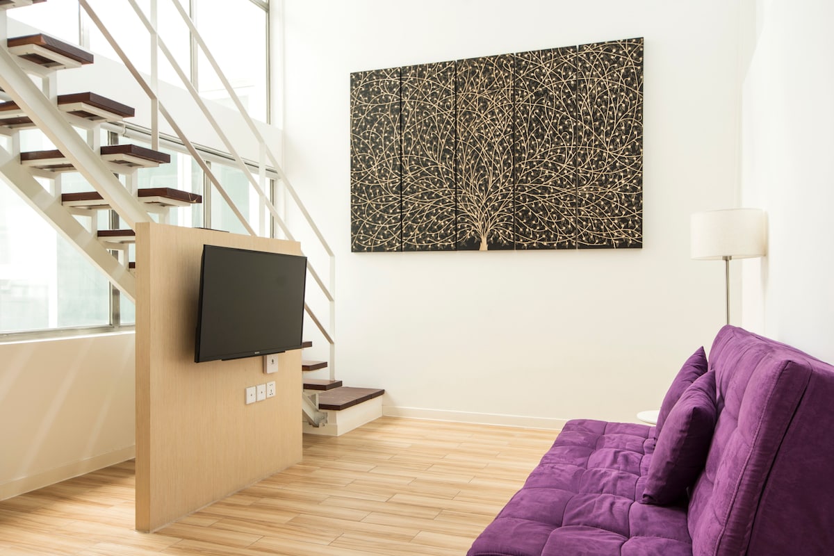 A bright living area features a purple sofa facing a mounted television. A large, textured wall art piece depicting a tree is displayed above the sofa. Natural light enters through large windows, highlighting the wooden flooring and stairway that leads to an upper level.