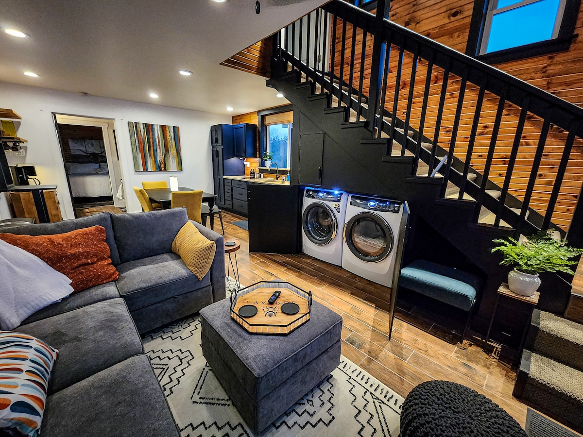 An inviting living space features a comfortable sectional sofa with decorative pillows. A sleek set of washer and dryer machines is visible beside a dining area. Dark wood accents and natural light from large windows enhance the overall ambiance of the open layout.