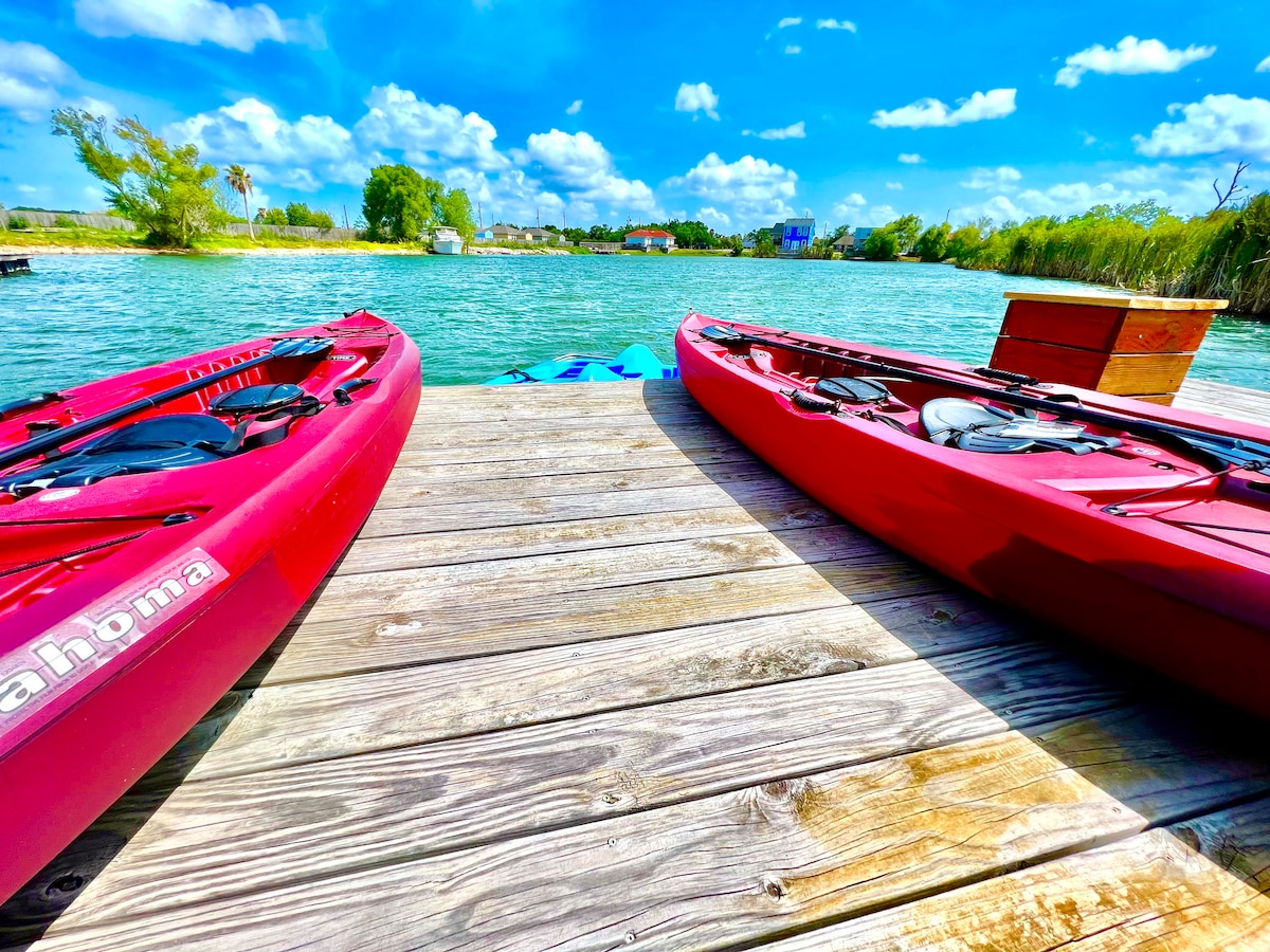 Two red kayaks rest on a wooden dock, positioned beside a calm lake. Lush greenery and distant buildings can be seen along the water's edge under a bright blue sky with fluffy white clouds.