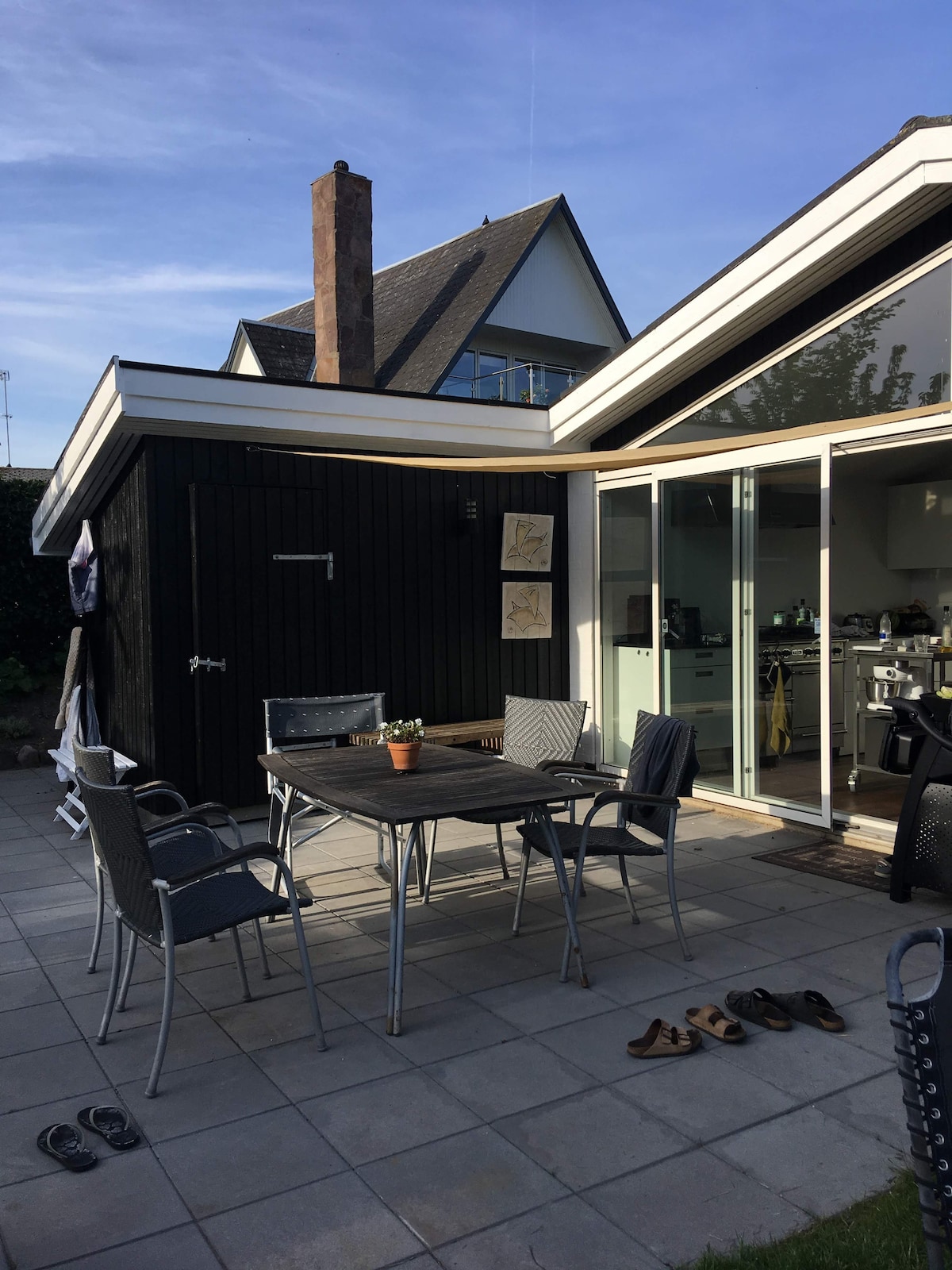 An outdoor terrace area is shown, featuring a wooden table surrounded by six chairs. A small potted plant is placed on the table. Sliding glass doors provide access to a modern kitchen inside, and a dark wooden shed is visible at the side.