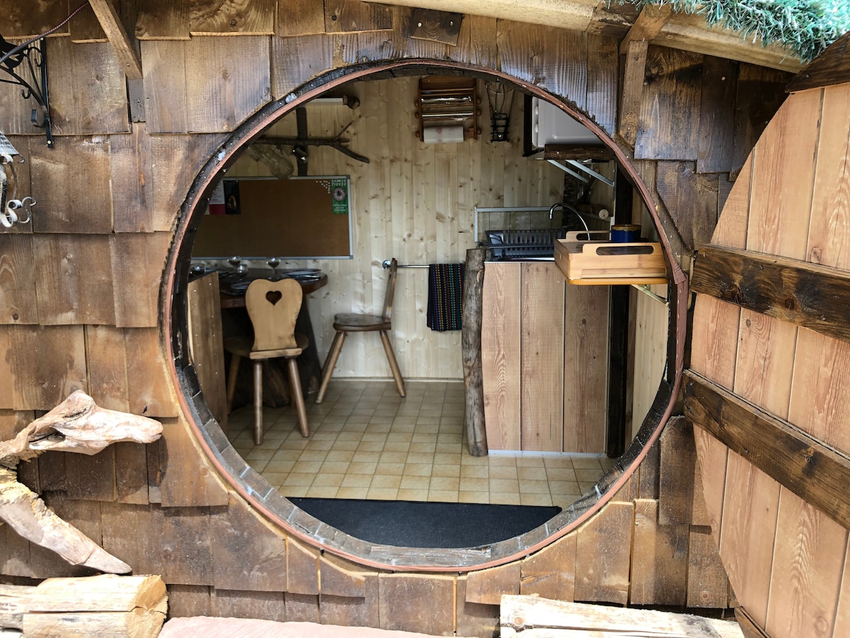 A round wooden door frames a view of the interior, where a compact kitchen and dining area are visible. Wooden countertops and chairs are highlighted, while the natural wood textures create an inviting environment. The space is illuminated by soft lighting, enhancing the cozy feel.