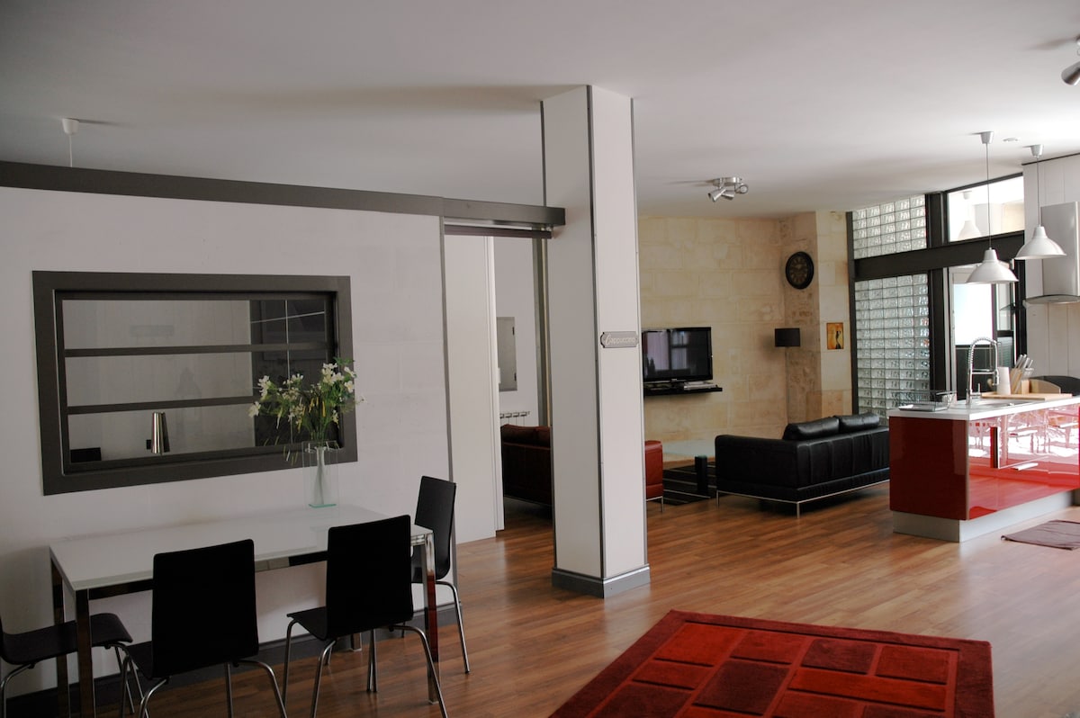 An open living area features modern furniture with a spacious black couch and sleek dining table. Large windows allow natural light to fill the room, highlighting the wood flooring. A decorative area rug provides a touch of warmth to the space.