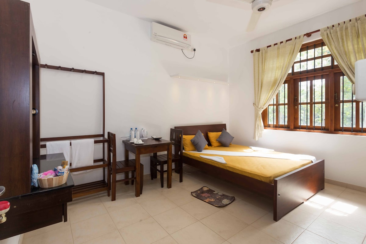 A well-lit room features a spacious bed with bright yellow bedding. A wooden table and chairs are positioned nearby. Large windows with wooden frames allow natural light in, while light curtains soften the ambiance. Amenities like water bottles and towels are visible.