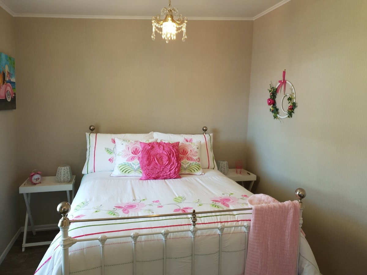 A cozy bedroom features a spacious bed with floral bedding and a decorative pink pillow. A small table is positioned on each side, with soft lighting from charming lamps. A decorative wreath is displayed on the wall, adding a touch of color to the neutral tones.