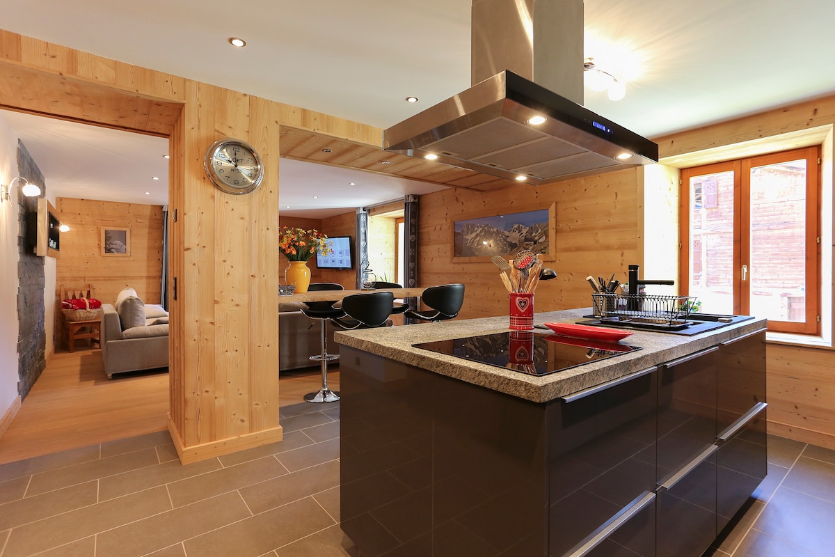 The kitchen features a central island with a dark countertop and modern appliances. Black bar stools surround the island, and wooden walls add a warm touch. Natural light enters through windows, illuminating the space. An open layout connects to the living area and dining space.