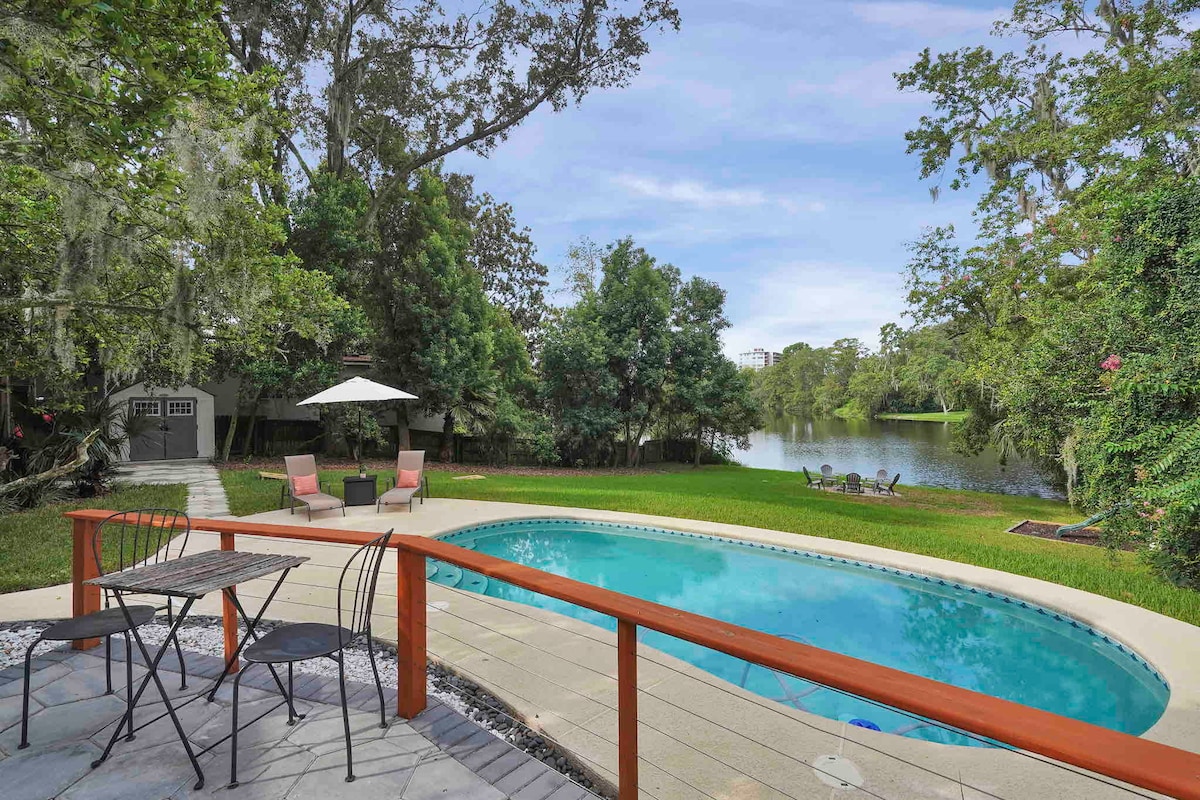 A serene pool area is situated beside a calm river, surrounded by lush greenery. Two lounge chairs are positioned on the wooden deck, with a parasol providing shade. A small table with two chairs is set nearby, inviting relaxation in the peaceful outdoor setting.