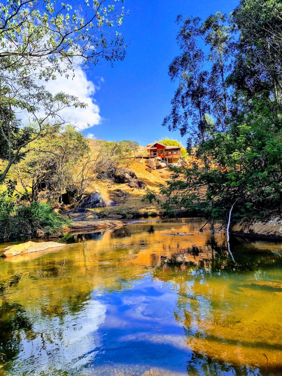 A serene river scene is captured, reflecting the clear blue sky and surrounding greenery. On the hillside, a newly built chalet is visible among the trees, with a harmonious blend of natural elements and peaceful water enhancing the tranquil ambiance.