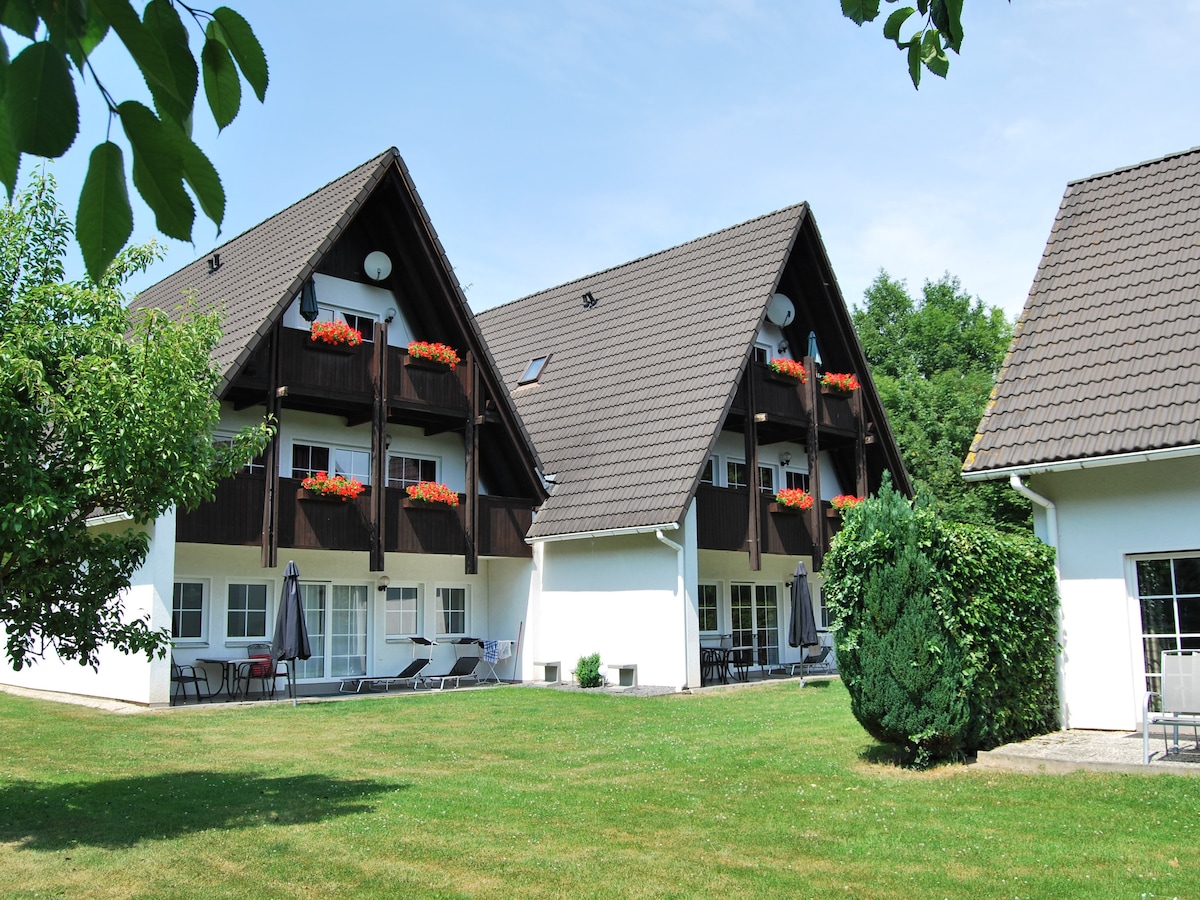 Two symmetrical buildings are depicted, showcasing a mix of dark wood and white façades. Bright flower boxes with vibrant flowers adorn the upper balconies, while shaded patio areas below feature seating arrangements. An expansive, well-manicured lawn stretches between the structures, framed by leafy trees.