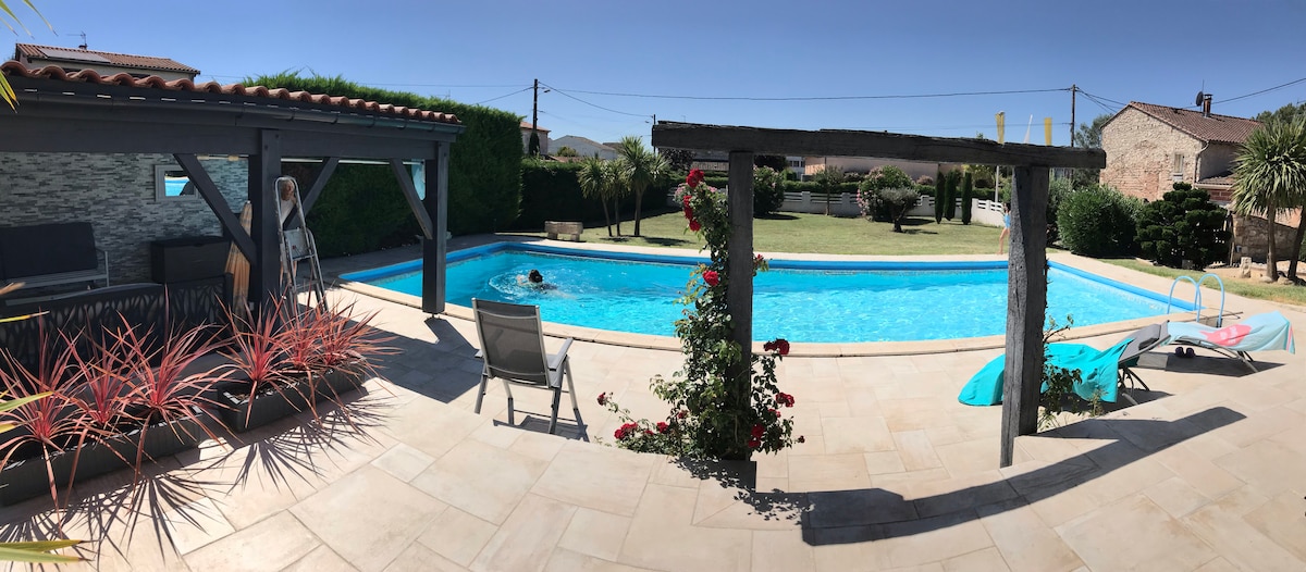 A spacious outdoor area features a large swimming pool surrounded by a grassy lawn. A wooden pergola with climbing plants provides shade, while lounge chairs invite relaxation. Bright blue skies complement the serene atmosphere, creating an inviting space for enjoyment.