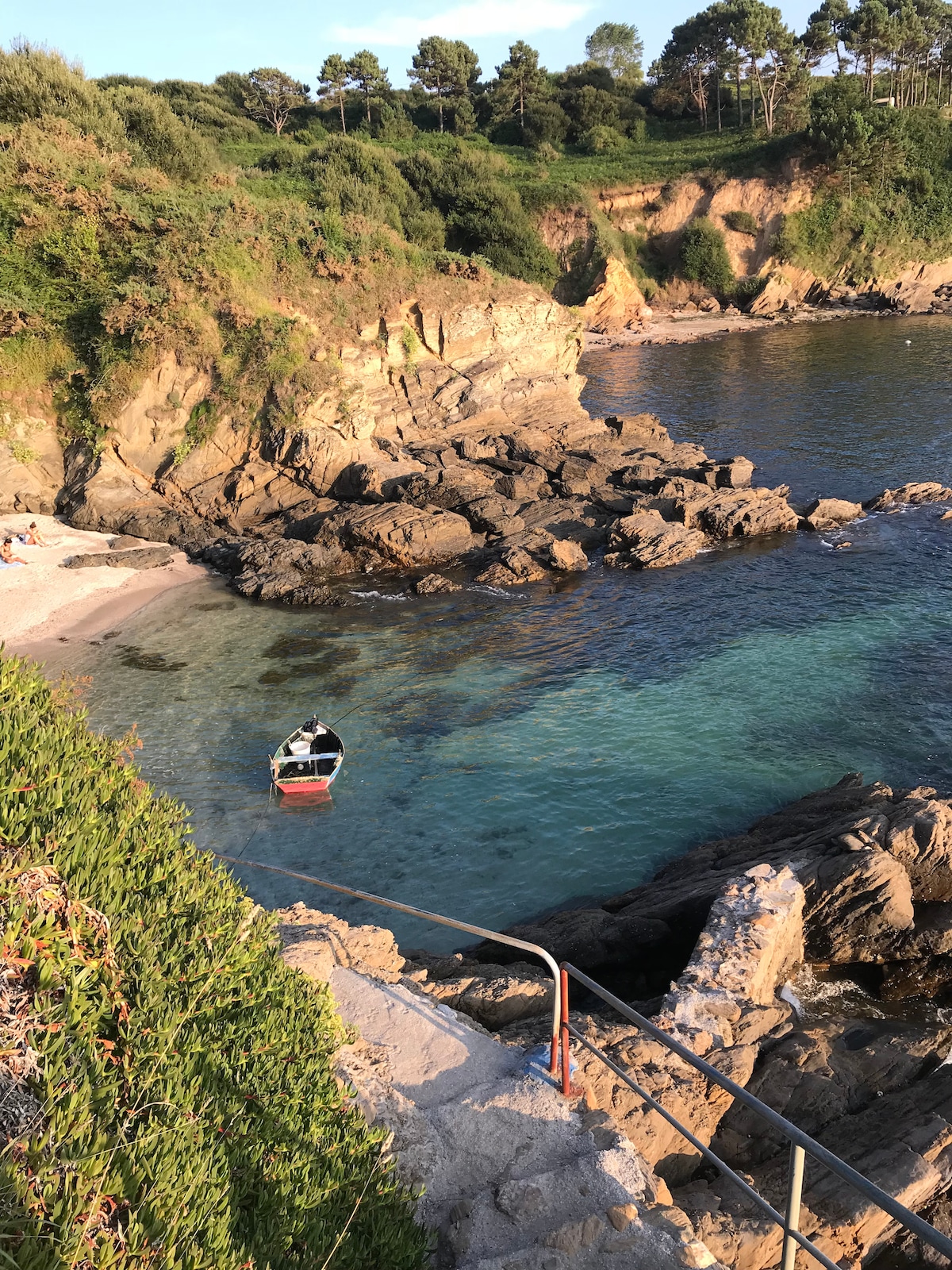 A coastal scene featuring rocky cliffs and a clear blue sea. A small boat is moored near a pebble beach, while people can be seen relaxing on the shore. Lush greenery surrounds the area, enhancing the natural beauty of the landscape.
