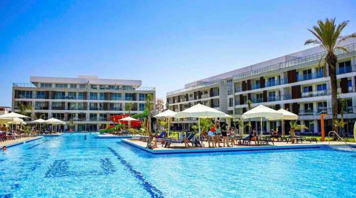 The image showcases a spacious pool area surrounded by modern buildings. Sun loungers and umbrellas line the water, offering shaded seating. Water slides and lush greenery are visible in the background, along with guests enjoying the sunshine, creating a lively atmosphere.