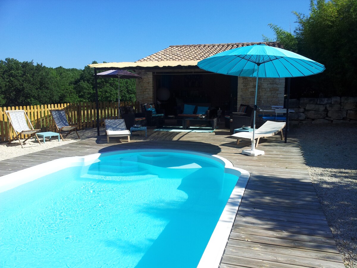 A sparkling swimming pool is framed by wooden decking, featuring lounge chairs for relaxation. A shaded seating area with an umbrella provides a cool retreat, while the surrounding greenery enhances the tranquil setting under a clear blue sky.