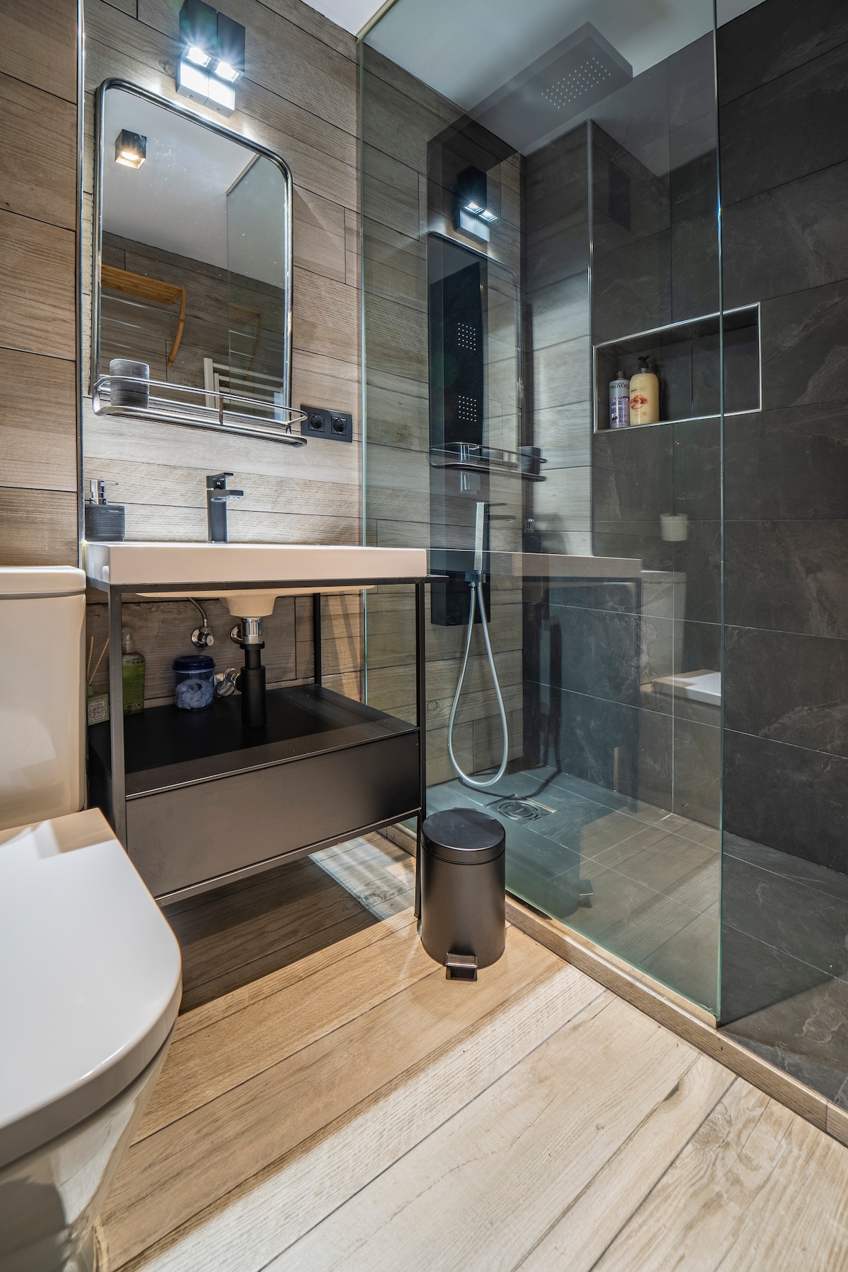 A modern bathroom is displayed, featuring a clean, glass-enclosed shower area and a compact vanity with storage beneath. Natural wood tones are complemented by sleek, dark tiles. Soft lighting enhances the atmosphere, and essential toiletries are neatly arranged on the shelves.
