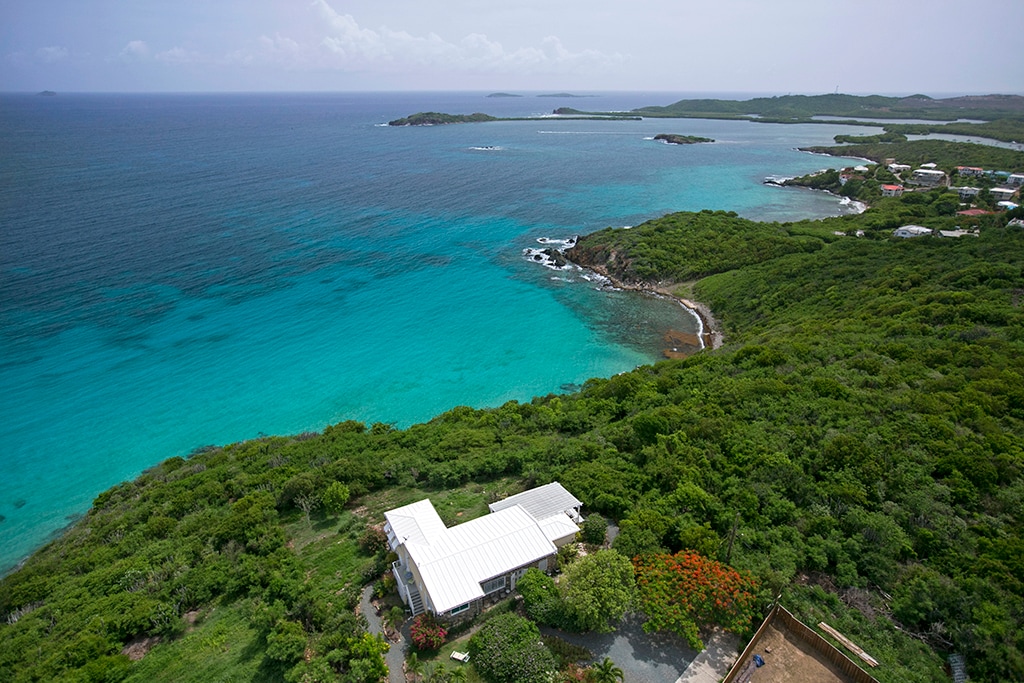 An aerial view highlights the Stonehouse property, nestled amid lush greenery along the coast. Crystal-clear turquoise waters stretch towards the horizon, dotted with small islands. The landscape transitions gently from vibrant vegetation to sandy beaches, emphasizing the serene beauty of the East End of St. Thomas.
