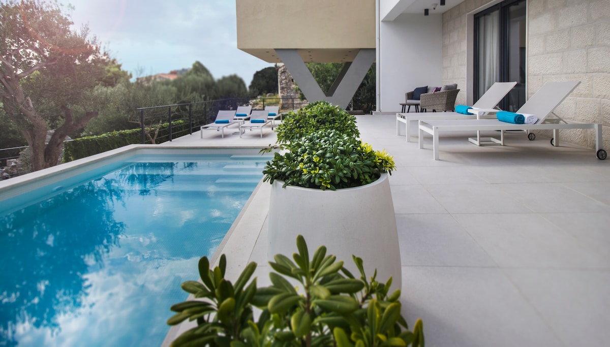 The outdoor area features a heated swimming pool bordered by modern lounge chairs and vibrant greenery. A spacious terrace, designed for relaxation, offers sun loungers and a tranquil ambiance complemented by the olive trees in the background.