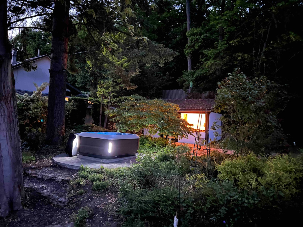 A tranquil outdoor area is shown, featuring a warm-lit building amidst lush greenery. A Jacuzzi is located on a stone platform, subtly illuminated by built-in lights. Surrounding trees and plants add to the peaceful and natural setting.