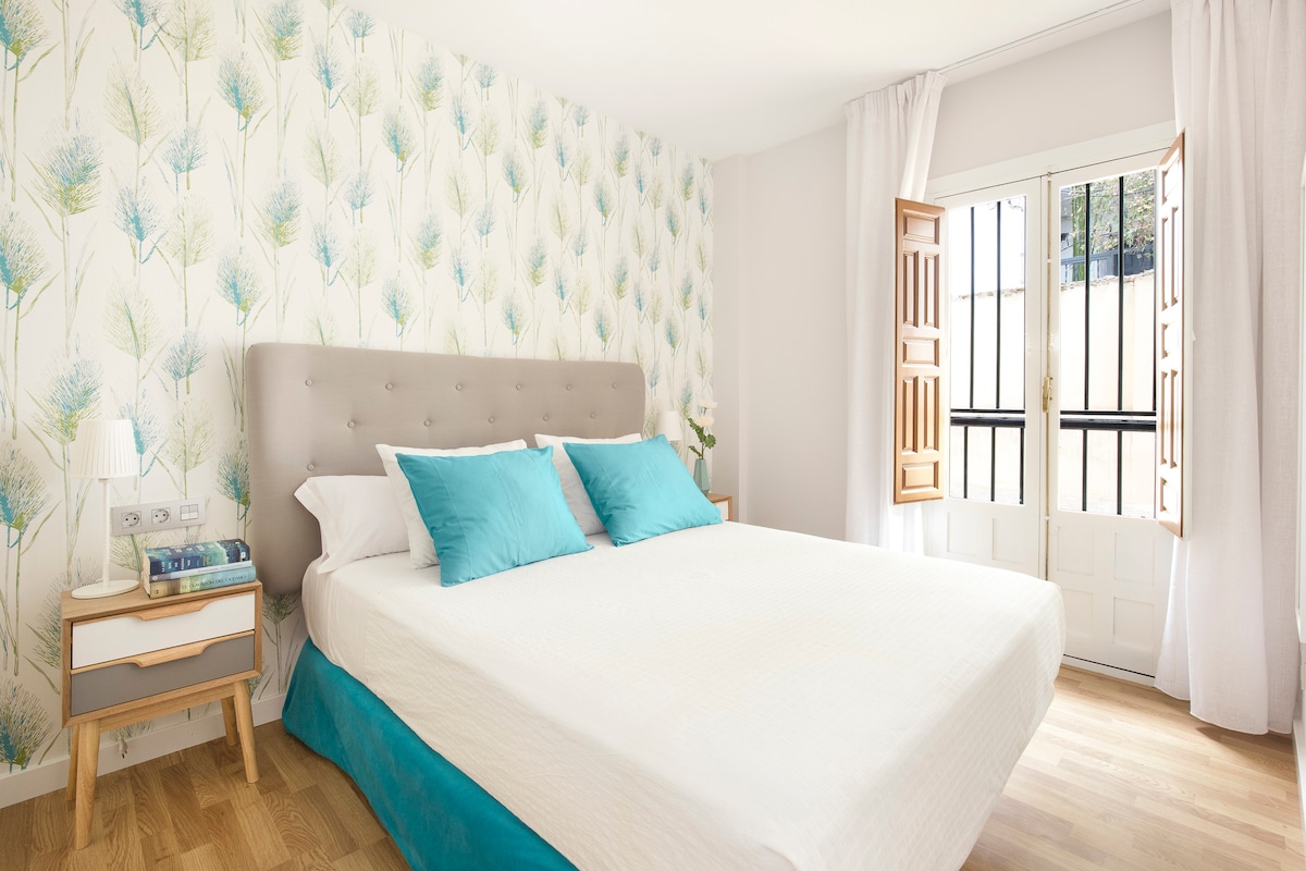 A spacious bedroom features a light-colored bed with decorative blue pillows. A vintage-style window opens to the outside, allowing natural light to fill the room. The wall displays a subtle botanical pattern, adding a touch of nature to the space, complemented by warm wooden flooring.