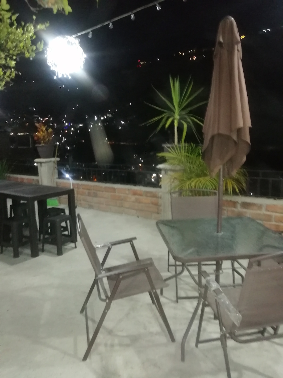 An outdoor terrace is shown under evening lights, featuring a glass table surrounded by four metal chairs. A large umbrella offers shade, while potted plants add greenery. A distant city view is visible, enhancing the serene atmosphere.