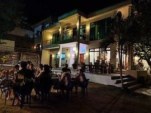 The resort features a two-story structure illuminated at night, showcasing large glass doors and windows. An outdoor area with seating is visible in the foreground, accompanied by guests enjoying a meal. Lush greenery surrounds the building, enhancing the welcoming atmosphere.