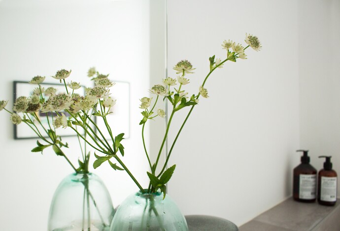 Two clear vases with fresh green flowers are displayed prominently. The background features a mirror reflecting light, while two bottles of liquid soap and lotion can be seen on a countertop, adding a touch of practicality.