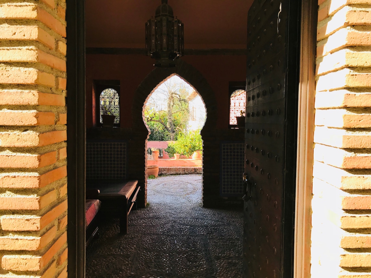 An inviting entrance is framed by an intricately designed archway, showcasing a glimpse of a lush garden visible through the open door. Soft light illuminates the textured stone floor, while traditional lanterns and wooden details enhance the welcoming atmosphere.