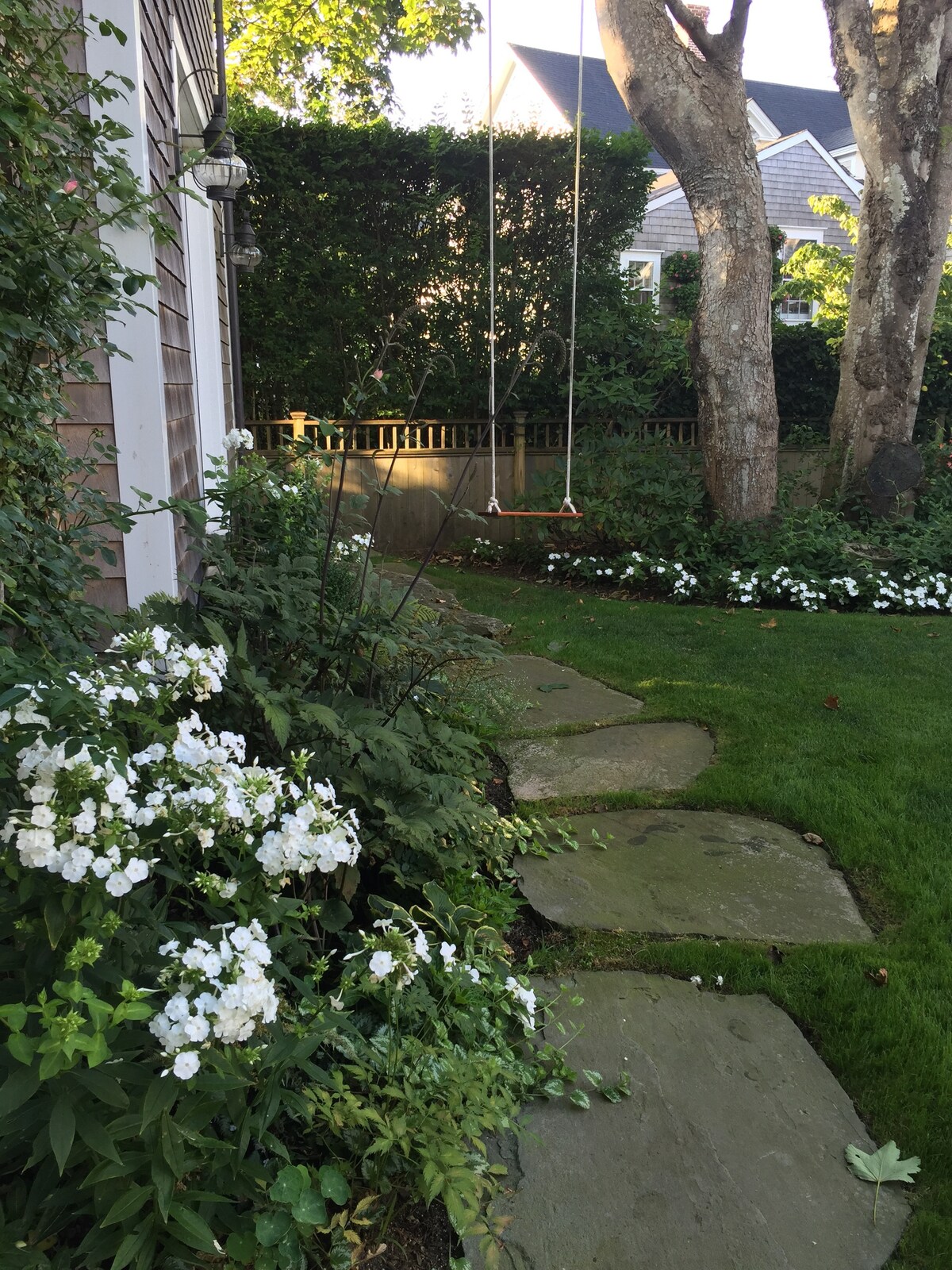 A garden path made of stone leads through a lush landscape featuring vibrant white flowers. A swing hangs from a tree nearby, adding a playful element to the serene outdoor space surrounded by greenery.