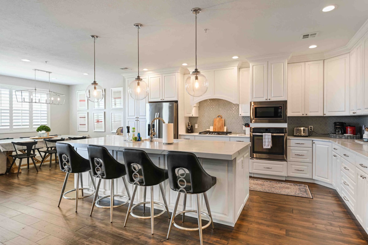 A modern kitchen features a large island with four stylish bar stools. White cabinetry complements stainless steel appliances, including a refrigerator, oven, and microwave. Pendant lights hang above the island, illuminating the open space, which is designed for both cooking and social gatherings.