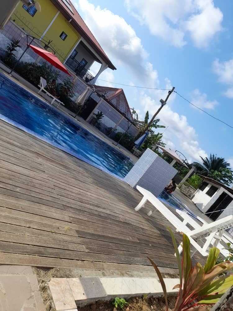 A spacious pool area is showcased, featuring a vibrant blue pool surrounded by wooden decking. A white lounge chair and red umbrella are positioned nearby, while various green plants add a touch of nature. The background reveals the colorful architecture of surrounding buildings under a bright sky.