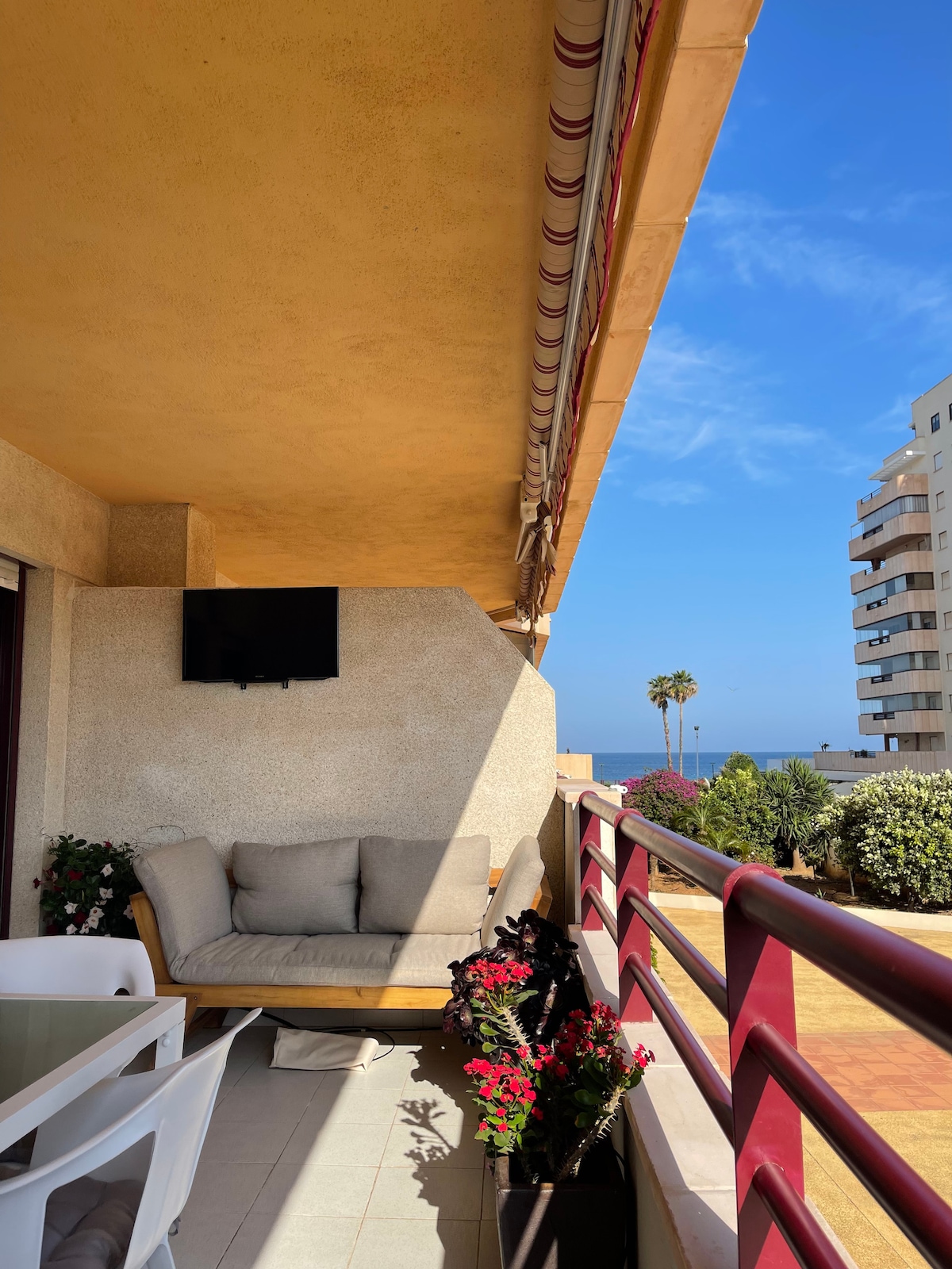 A cozy balcony is depicted, featuring a comfortable seating area with soft cushions against a backdrop of blue skies. Potted flowers add natural color, and a wall-mounted television is installed. The view includes nearby palm trees and the ocean in the distance.